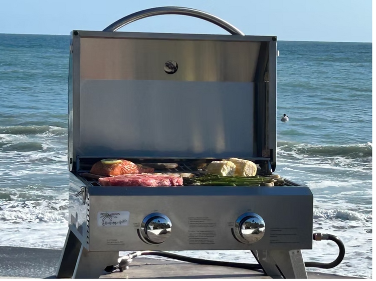 KoKoMo Portable Grill stainless steel tabletop BBQ grilling steak and vegetables on the beach with ocean background