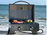 KoKoMo Portable Grill stainless steel tabletop BBQ grilling steak and vegetables on the beach with ocean background